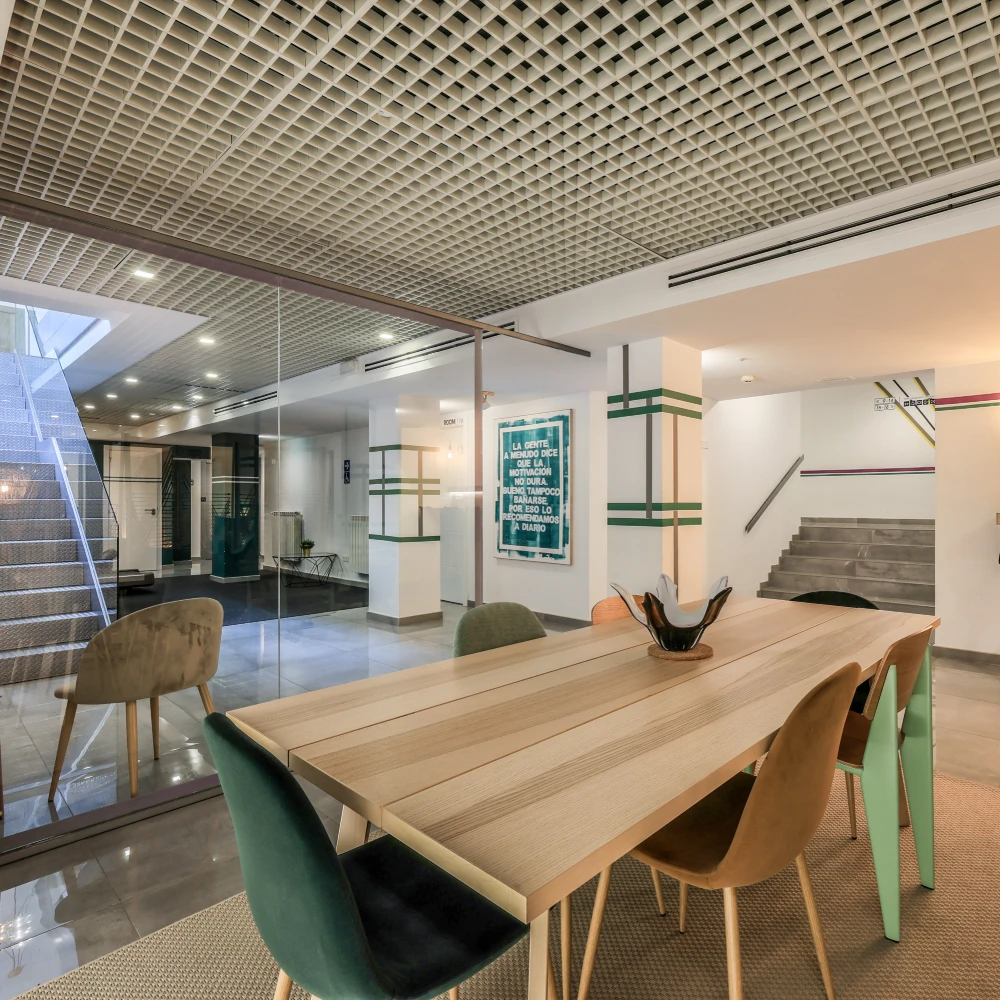 Sala de recepci&oacute;n con mesa de madera, sillas y escalera al fondo.