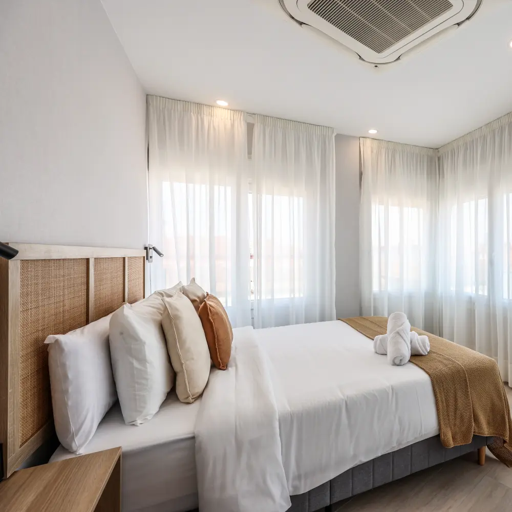 Dormitorio moderno con cama doble, cojines y cortinas blancas al sol