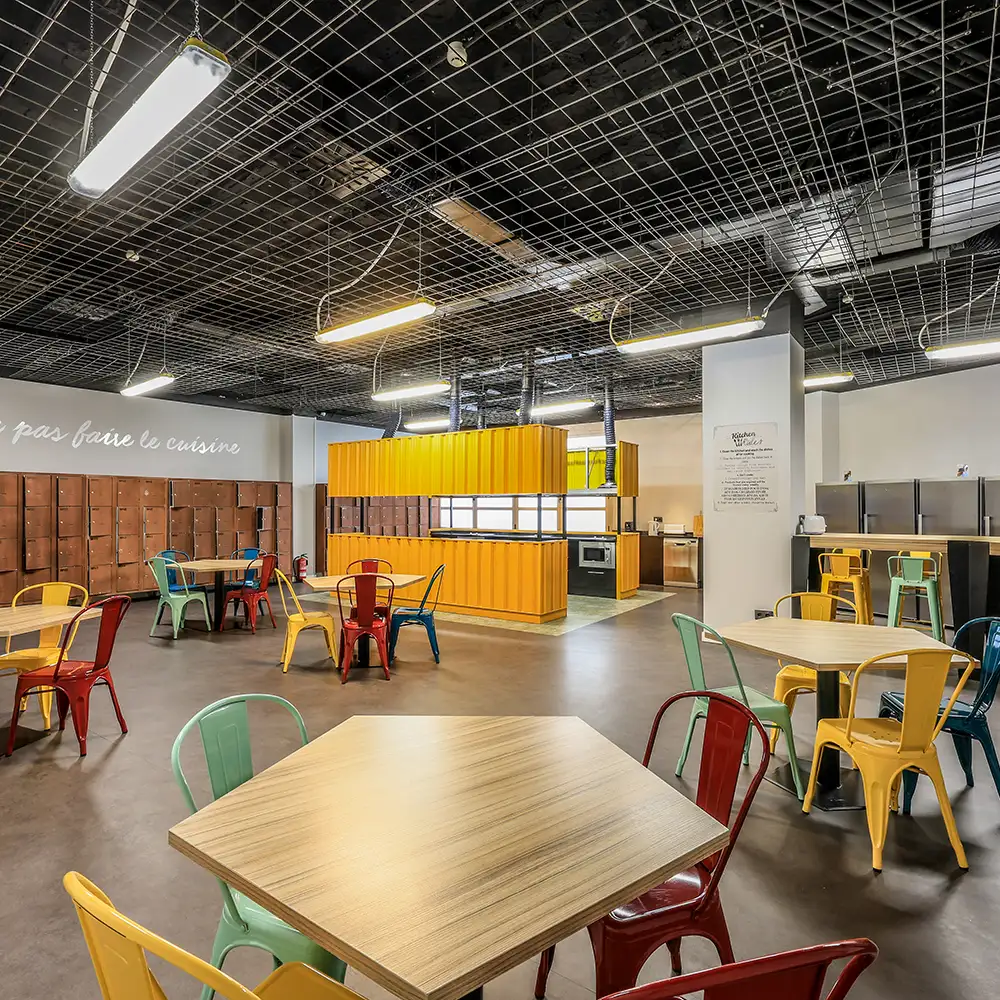 Comedor comunitario con mesas de madera, sillas de colores y cocina al fondo