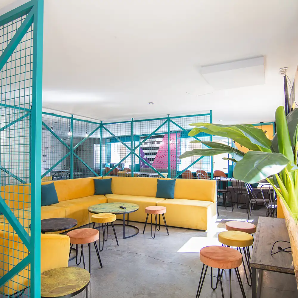 Interior de sala con sof&aacute; amarillo, mesas y sillas, plantas y rejas met&aacute;licas