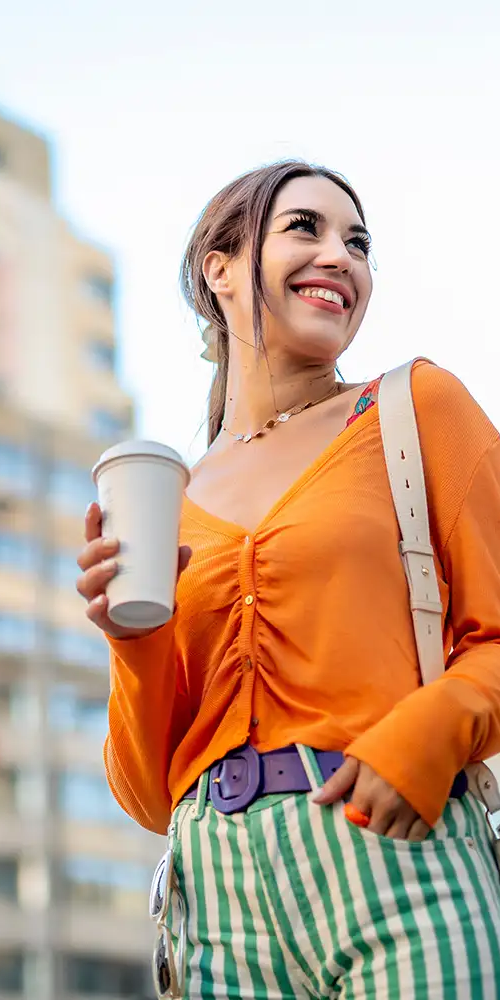 Mujer sonriente con taza de caf&eacute; en la calle, con blusa naranja y short verde