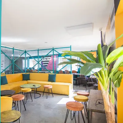 Terraza con mesas y sillas, sof&aacute;s amarillos y plantas tropicales frente a un parque infantil