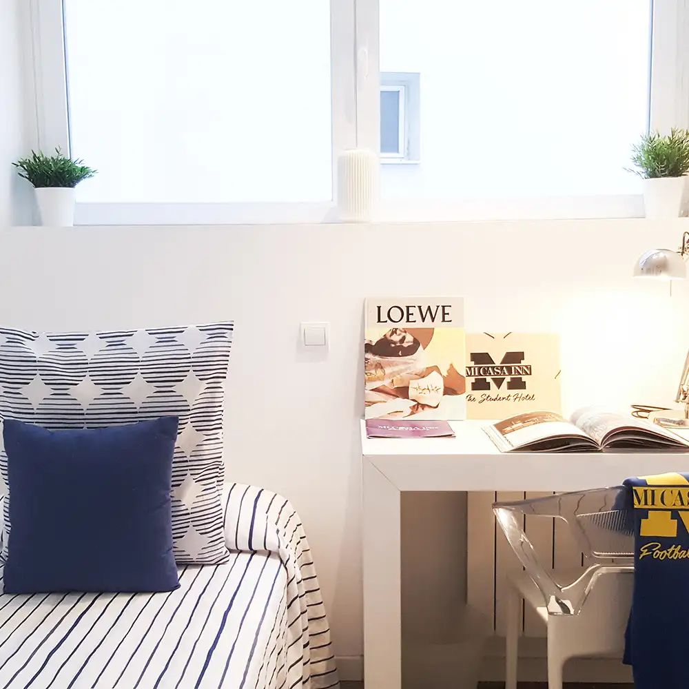 Dormitorio blanco con cama, cojines azules y escritorio con revista y l&aacute;mpara