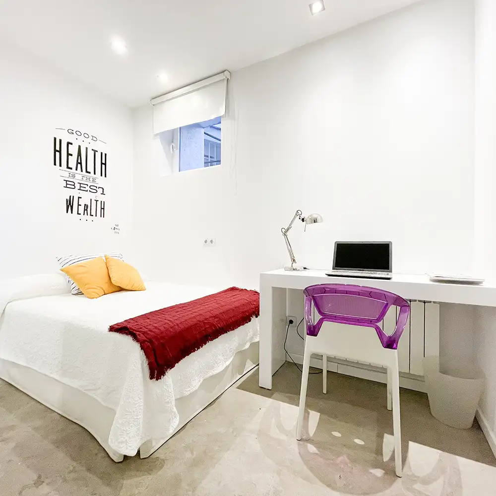 Habitaci&oacute;n blanca con cama, escritorio, laptop y silla morada transparente