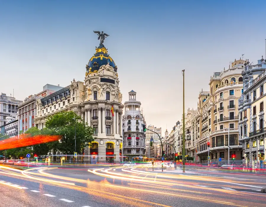Plaza urbana con estatua dorada y edificios hist&oacute;ricos al atardecer, luces de tr&aacute;fico