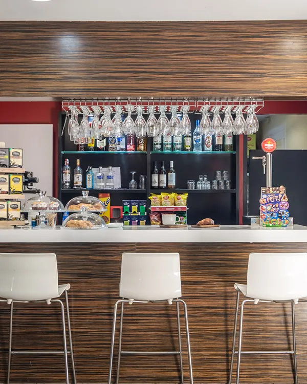 Bar con botellas, vasos y botellas de agua, interior con taburetes blancos.