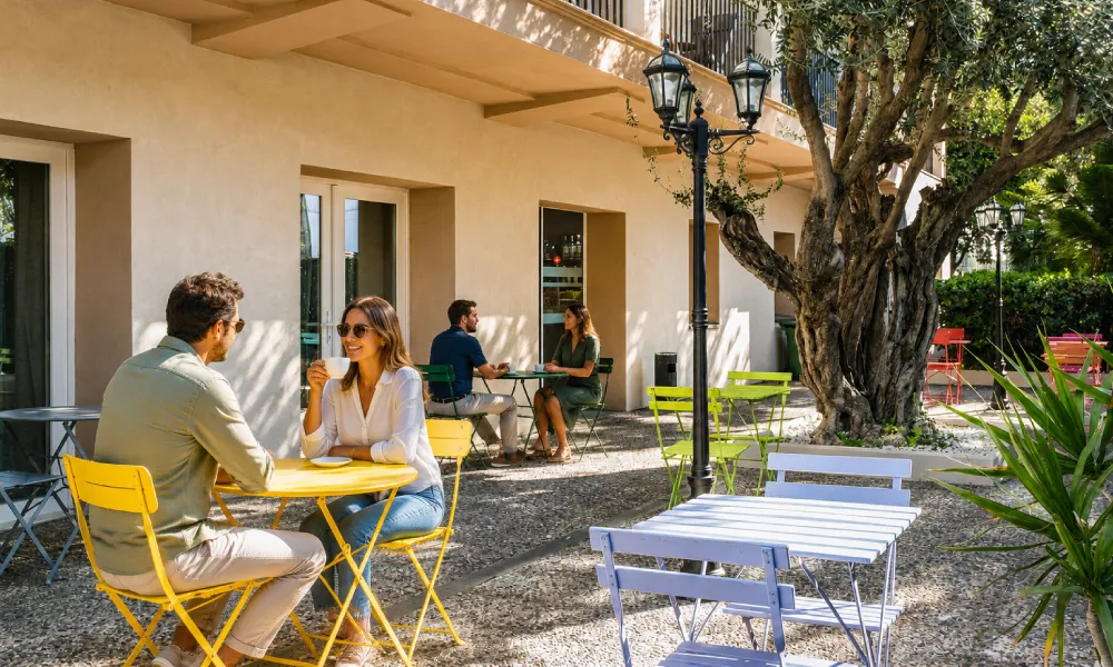 Personas conversan en patio con mesas, sillas de colores y &aacute;rbol grande.