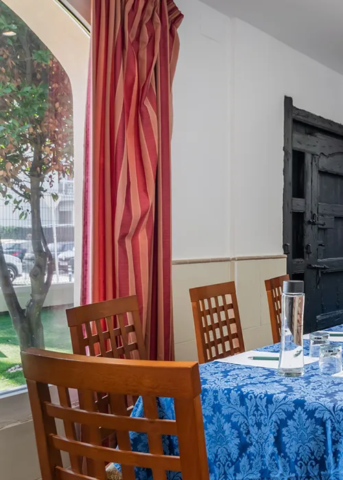 Mesa con mantel azul y sillas junto a cortinas rojas en interior de sal&oacute;n.