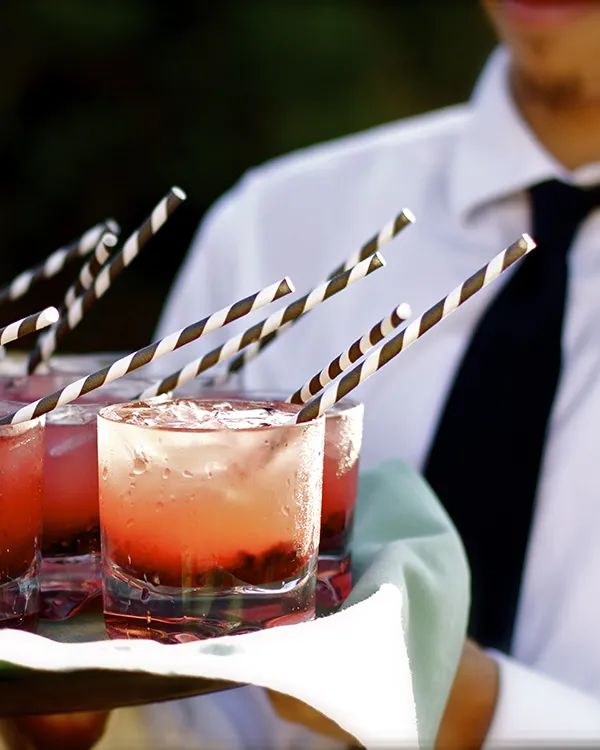 Bebidas rojas con hielo y pajillas rayadas, con camarero al fondo.