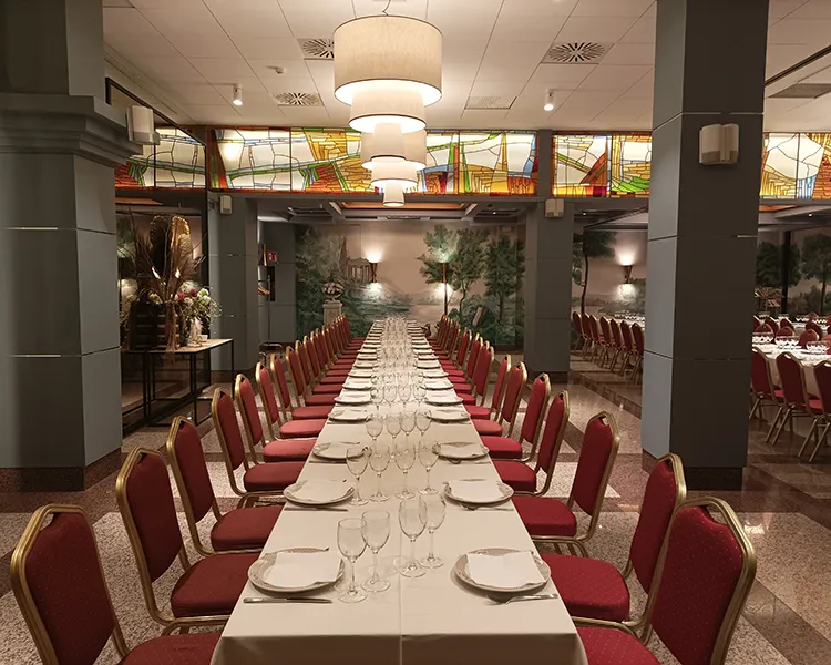 Sala de banquete con mesas servidas y sillas rojas, vitrales al fondo.