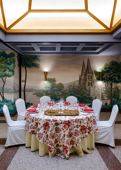 Mesa decorada con mantel floral y sillas blancas, fondo mural de iglesia.