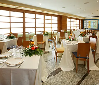 Sal&oacute;n de banquetes con mesas decoradas y sillas, ventanas grandes al fondo.