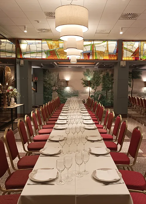 Mesa con cubiertos y copas, sal&oacute;n con sillas rojas y vitral al fondo.