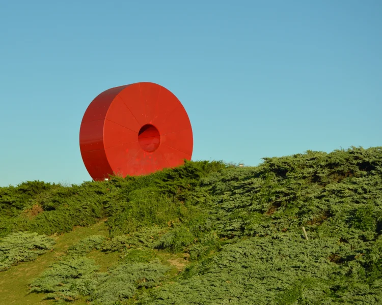 Escultura roja sobre una colina verde bajo un cielo azul