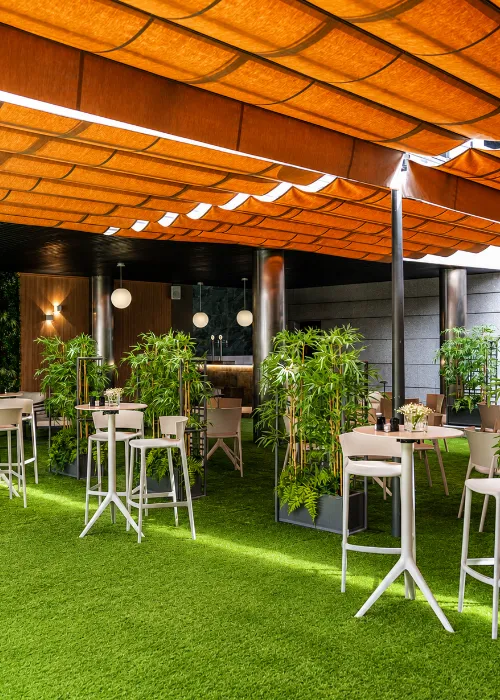 Terraza con c&eacute;sped, mesas y sillas blancas bajo carpa naranja con plantas