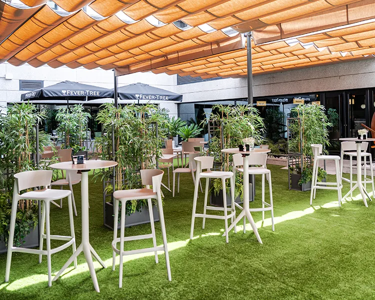 Terraza con c&eacute;sped y mesas blancas junto a plantas bajo un techo de madera.