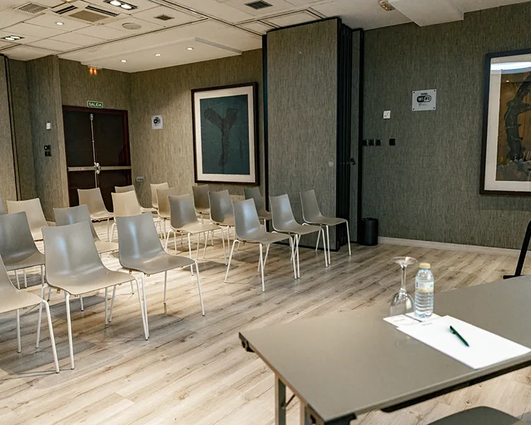 Sala de espera con sillas blancas, mesa y paredes grises, con obras enmarcadas