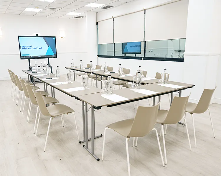 Sala de formaci&oacute;n con mesa rectangular, sillas blancas y pantallas de proyecci&oacute;n
