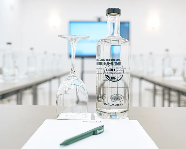 Botella de agua en mesa blanca con copa de cristal al fondo.