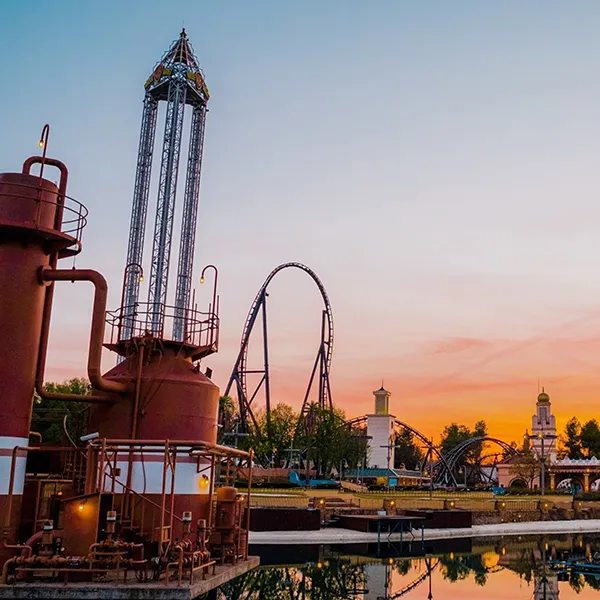 Parque tem&aacute;tico al atardecer con noria y torre industrial junto a lago.