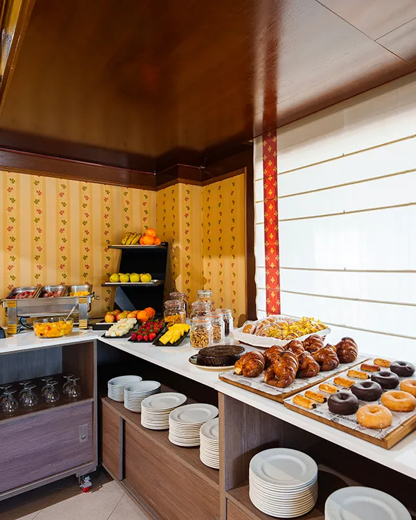 Buffet con reposter&iacute;a y bandejas, bebidas y frutas en una sala de desayuno.