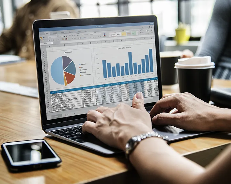 Persona trabajando en laptop con gr&aacute;ficos y tabla financiera, caf&eacute; al lado