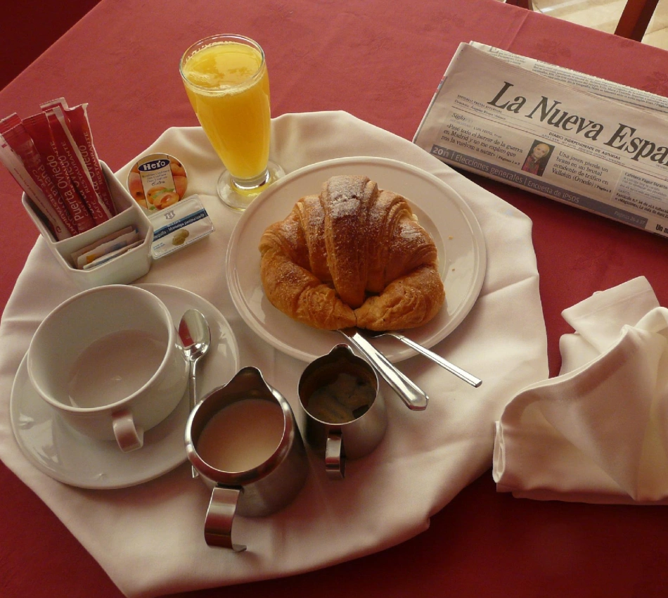 Desayuno en mesa con croissant, jugo y peri&oacute;dicos, sobre mantel rojo.