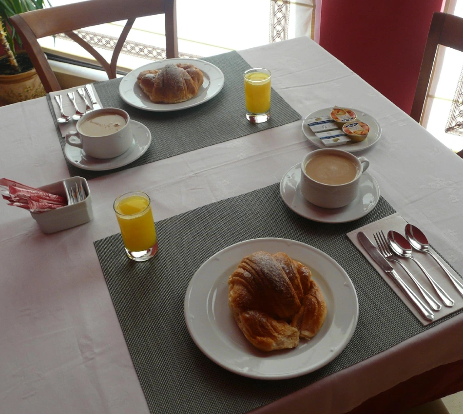 Mesa de desayuno con croissant, caf&eacute;, jugo y utensilios en un hotel.