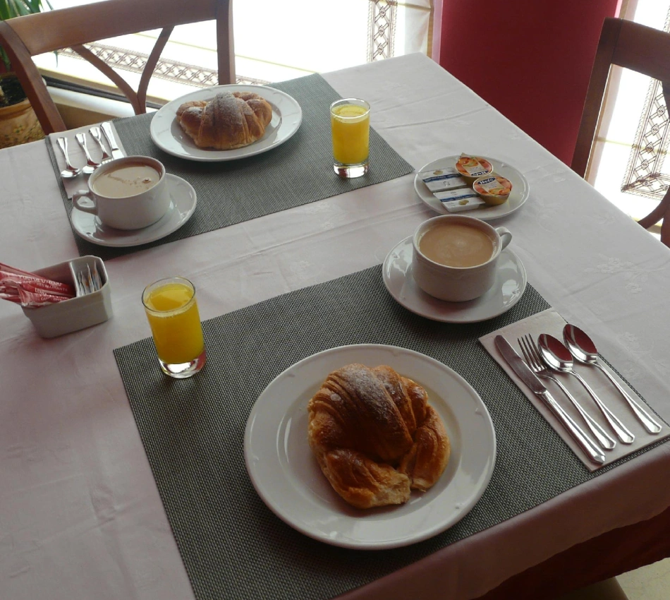 Mesa de desayuno con croissant, caf&eacute;, zumo y vajilla, en un comedor.