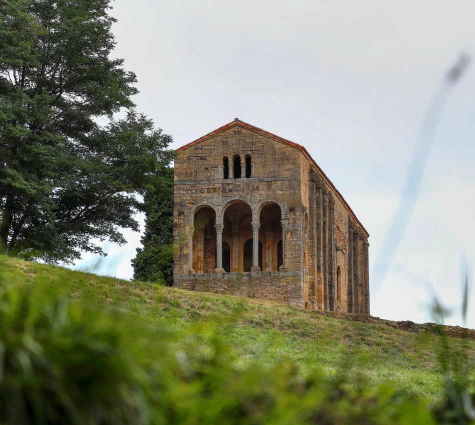 Iglesia rom&aacute;nica de piedra con arcos en ladera verde y cielo nublado