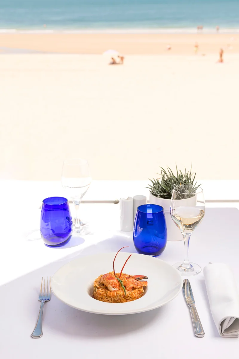 Mesa en la playa con plato de comida, vasos azules y copa de vino, vista al mar
