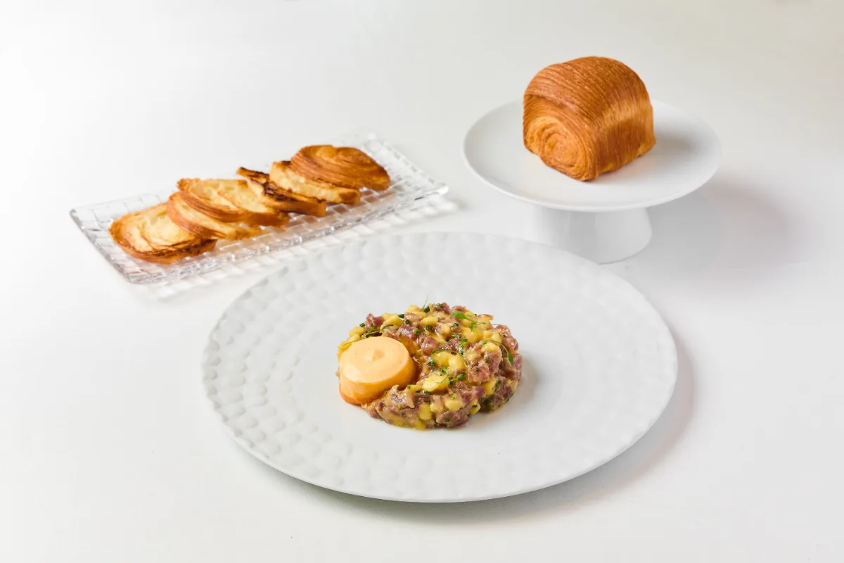 Desayuno servido: croissant, tocino, huevo y salchicha en plato blanco.