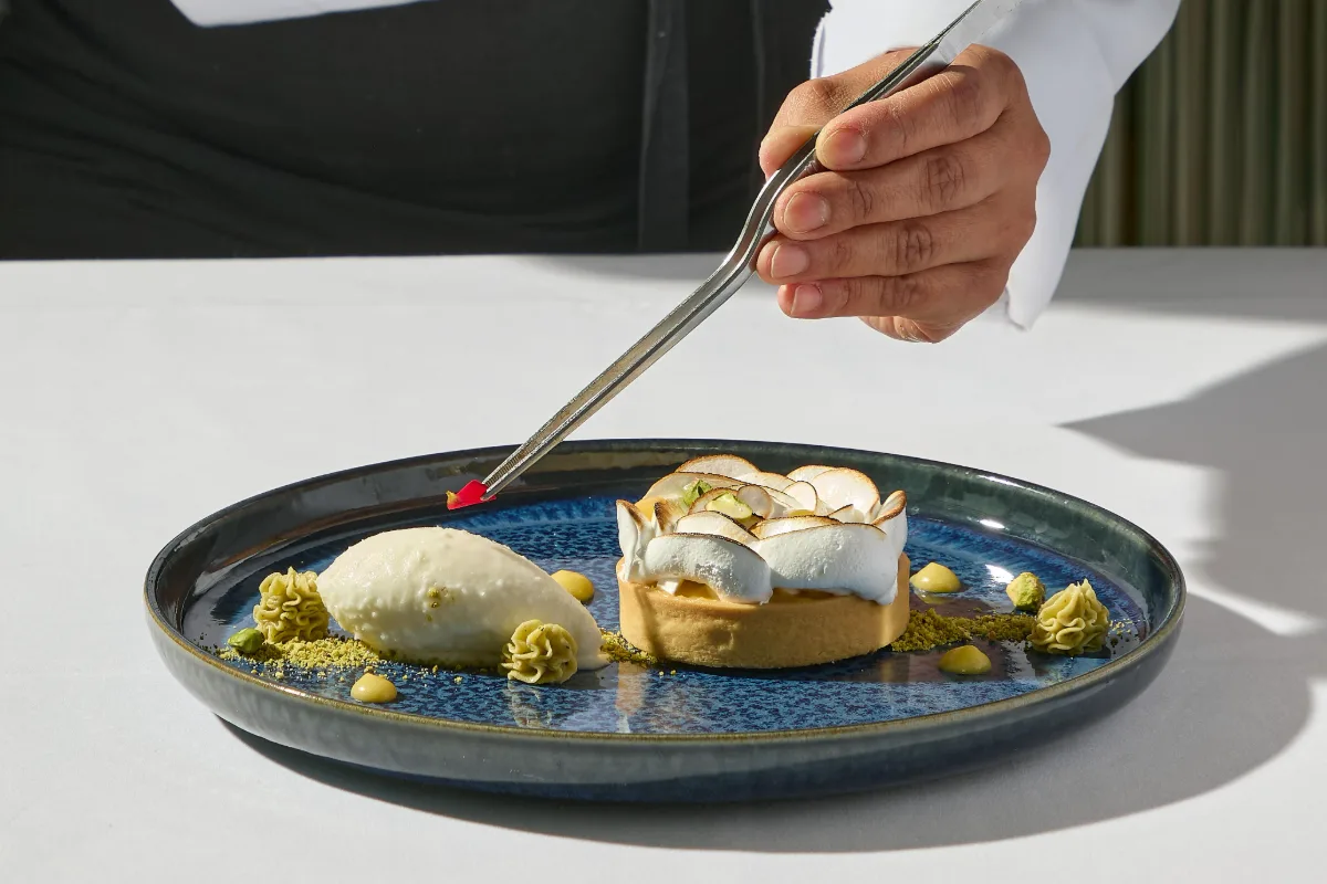 Mano con pinzas sirve un postre en plato azul con salsa y frutas