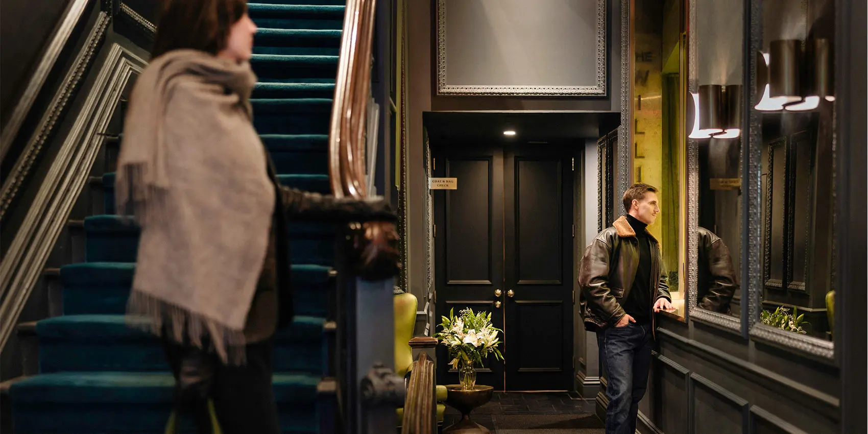 Man stands by doors in ornate hallway, framed by mirrors and teal stairs