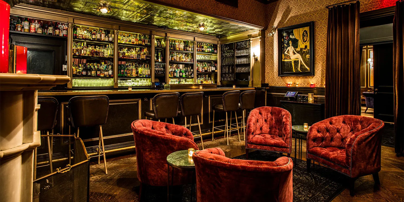 Warmly lit bar interior with backlit shelves, stools, and red velvet seating