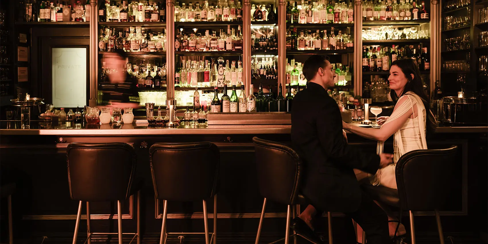 Bartender and customer seated at a warmly lit bar with many bottles