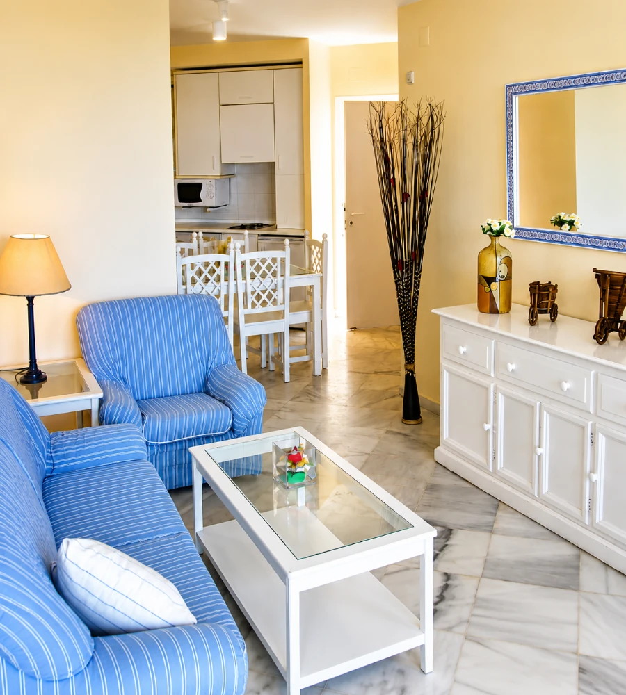 Sala de estar con sillones azules, mesa blanca y cocina al fondo