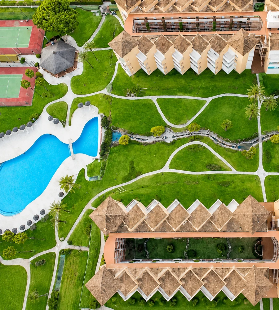 Vista a&eacute;rea de jardines verdes, piscina azul y edificios con tejados marrones.