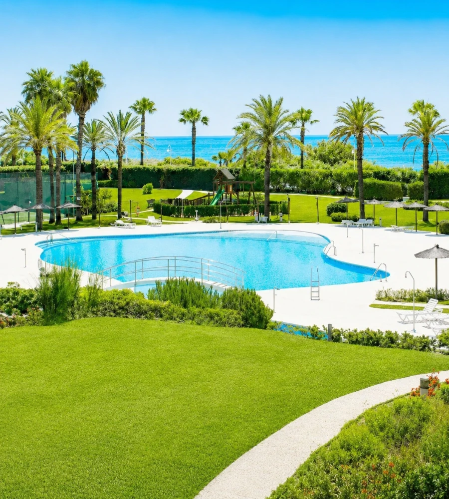 Piscina azul con palmeras y c&eacute;sped, vista al mar.