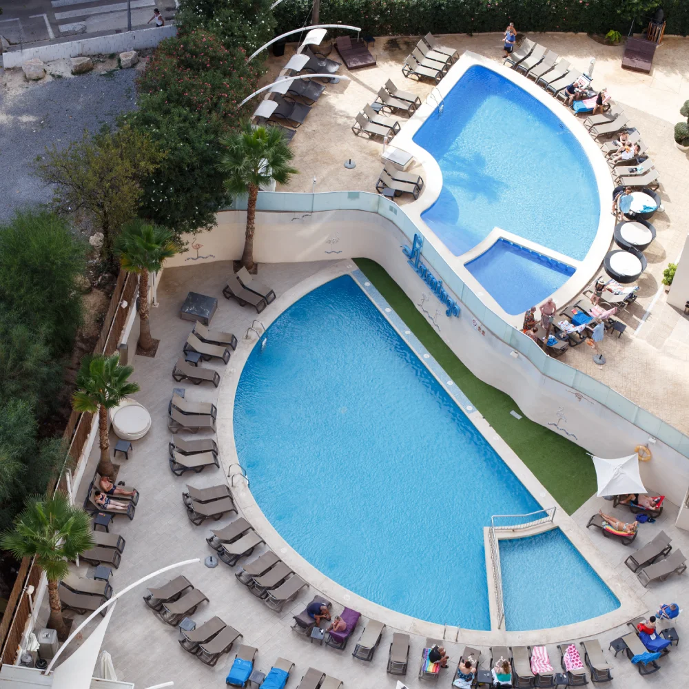 Vista a&eacute;rea de dos piscinas azules con tumbonas y hu&eacute;spedes en un hotel.