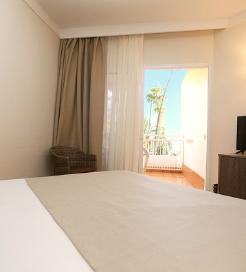 Habitaci&oacute;n con cama doble, cortinas y balc&oacute;n con palmeras al exterior.