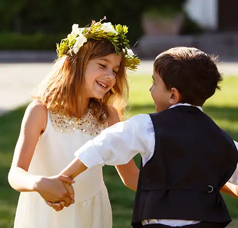 Ni&ntilde;a y ni&ntilde;o felices con coronas de flores, tom&aacute;ndose las manos al aire libre