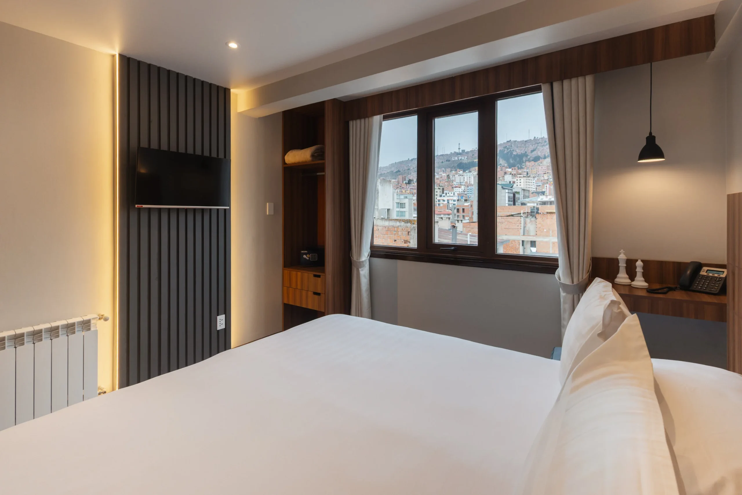 Dormitorio moderno con cama, TV y vista urbana por ventanas.