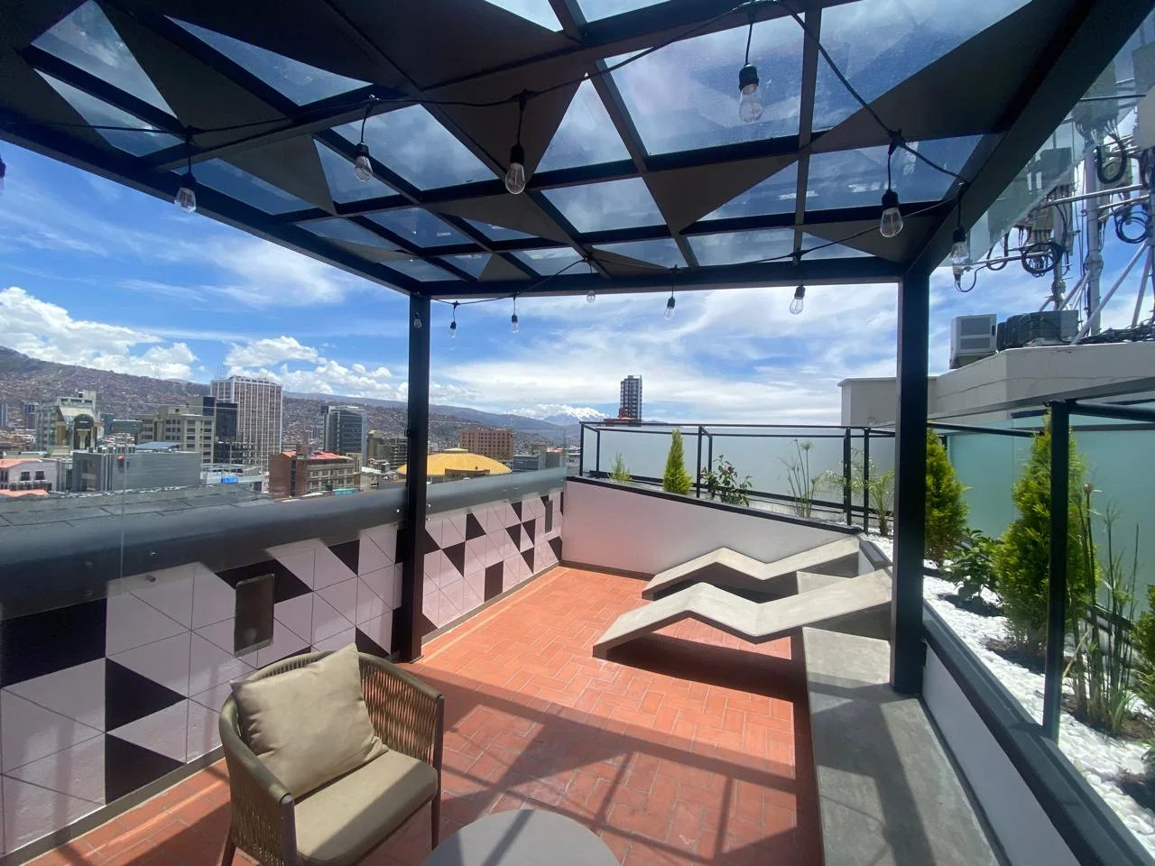 Terraza moderna con piso rojo, plantas y l&aacute;mparas bajo techo, vista urbana