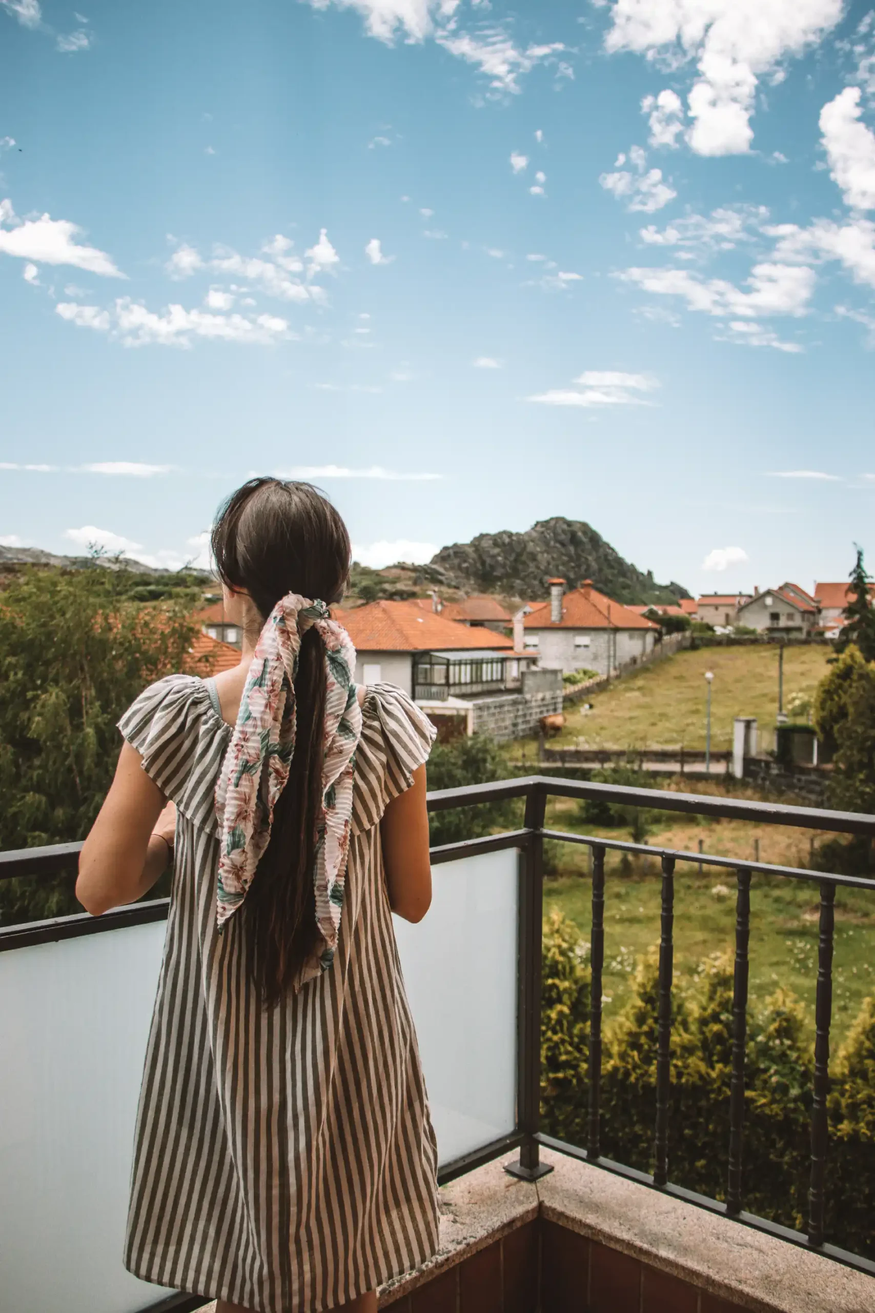 Menina de costas em varanda, olhando casas e montanhas sob c&eacute;u azul com nuvens