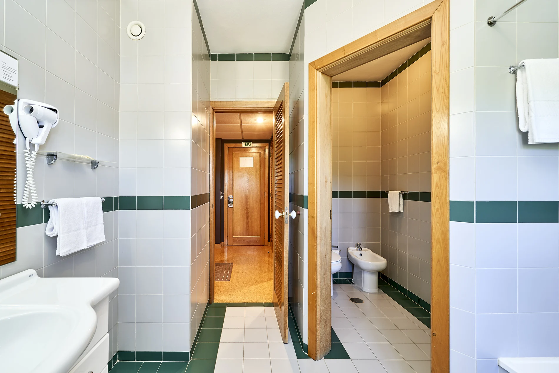 Corredor de banheiro com azulejos brancos e verdes, pia e toalhas.