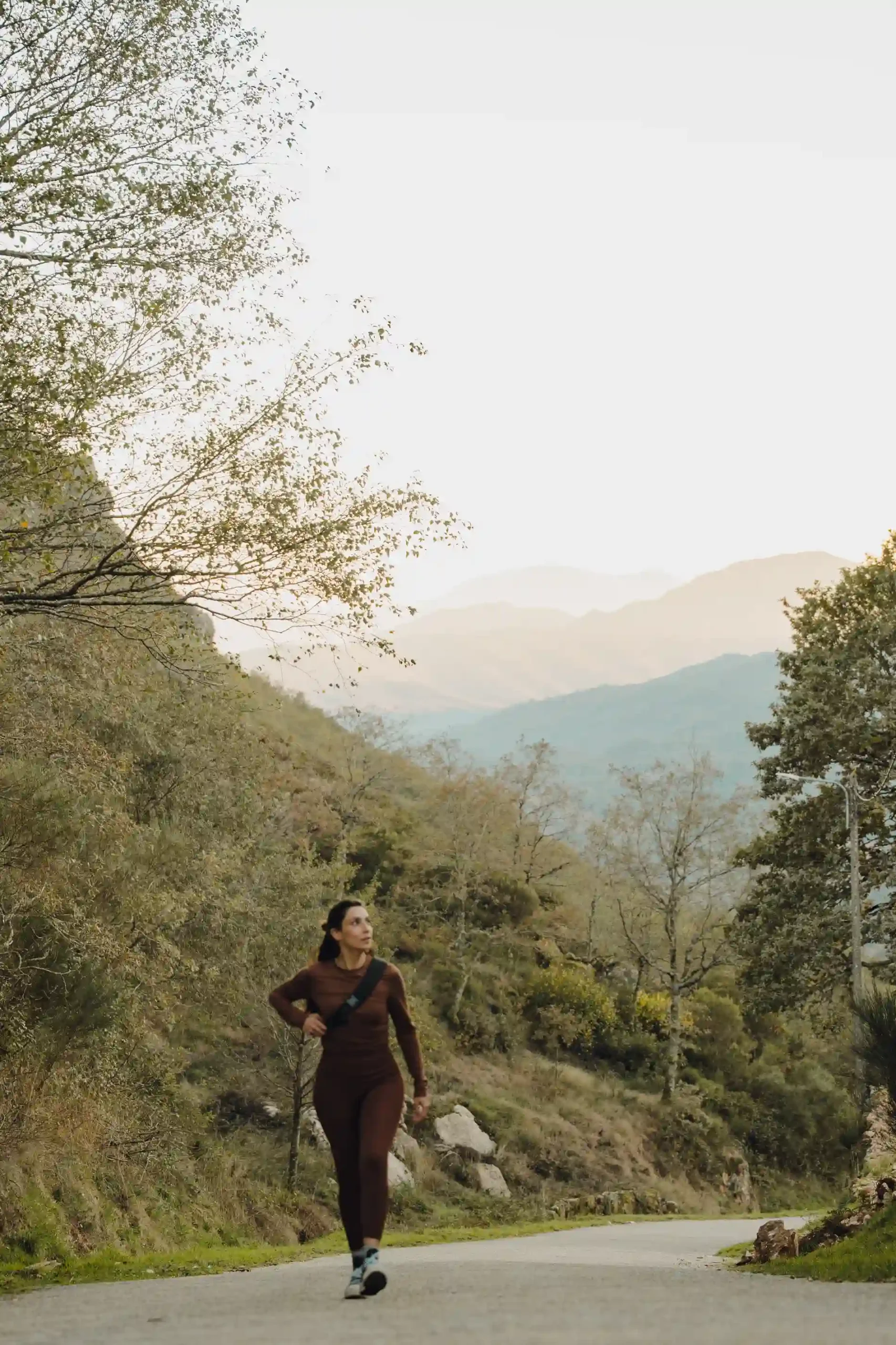 Mulher caminhando na trilha em meio a montanhas e vegetação ao entardecer