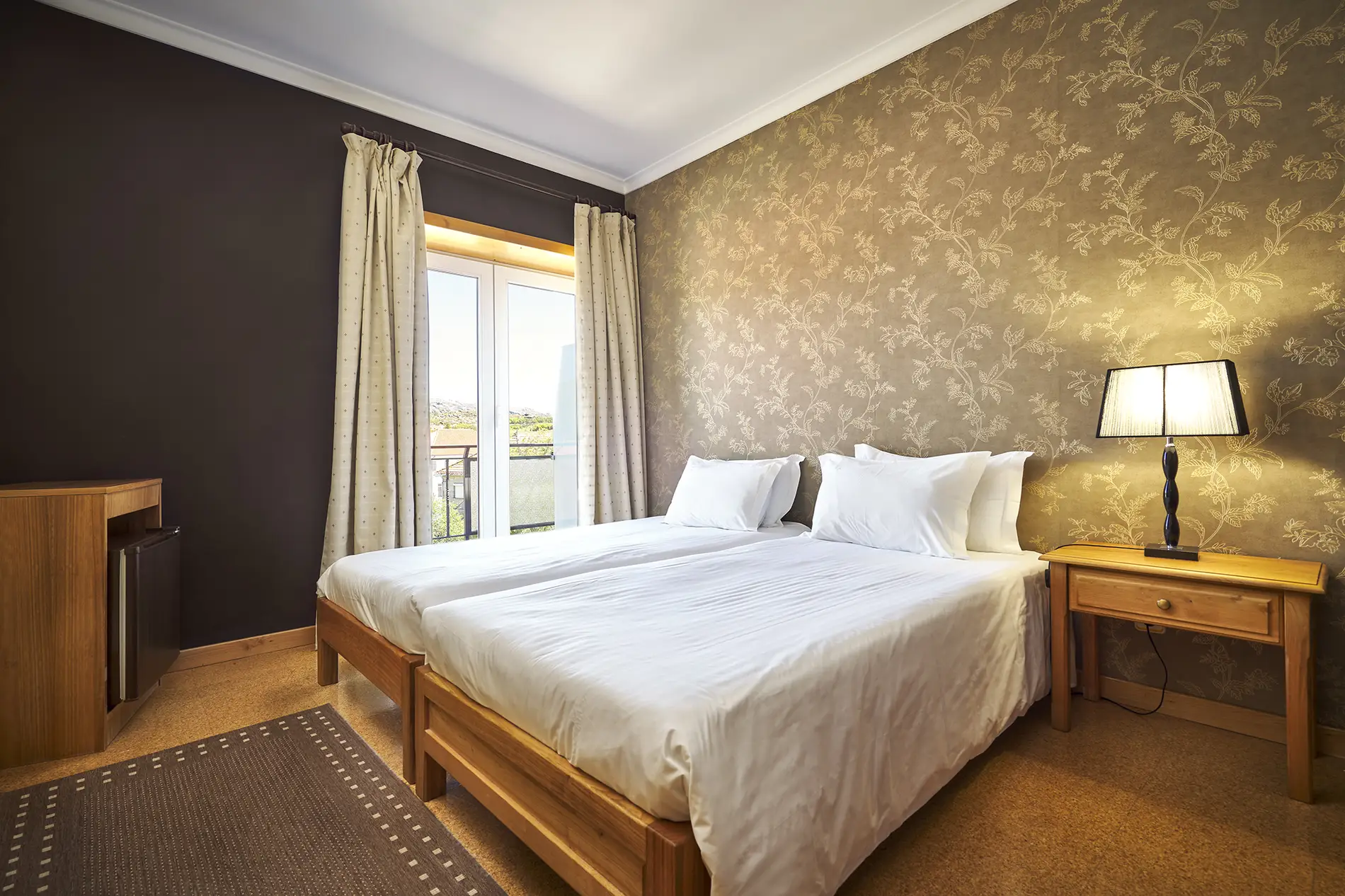 Quarto com cama de casal, roupa branca, papel de parede floral e cortinas.
