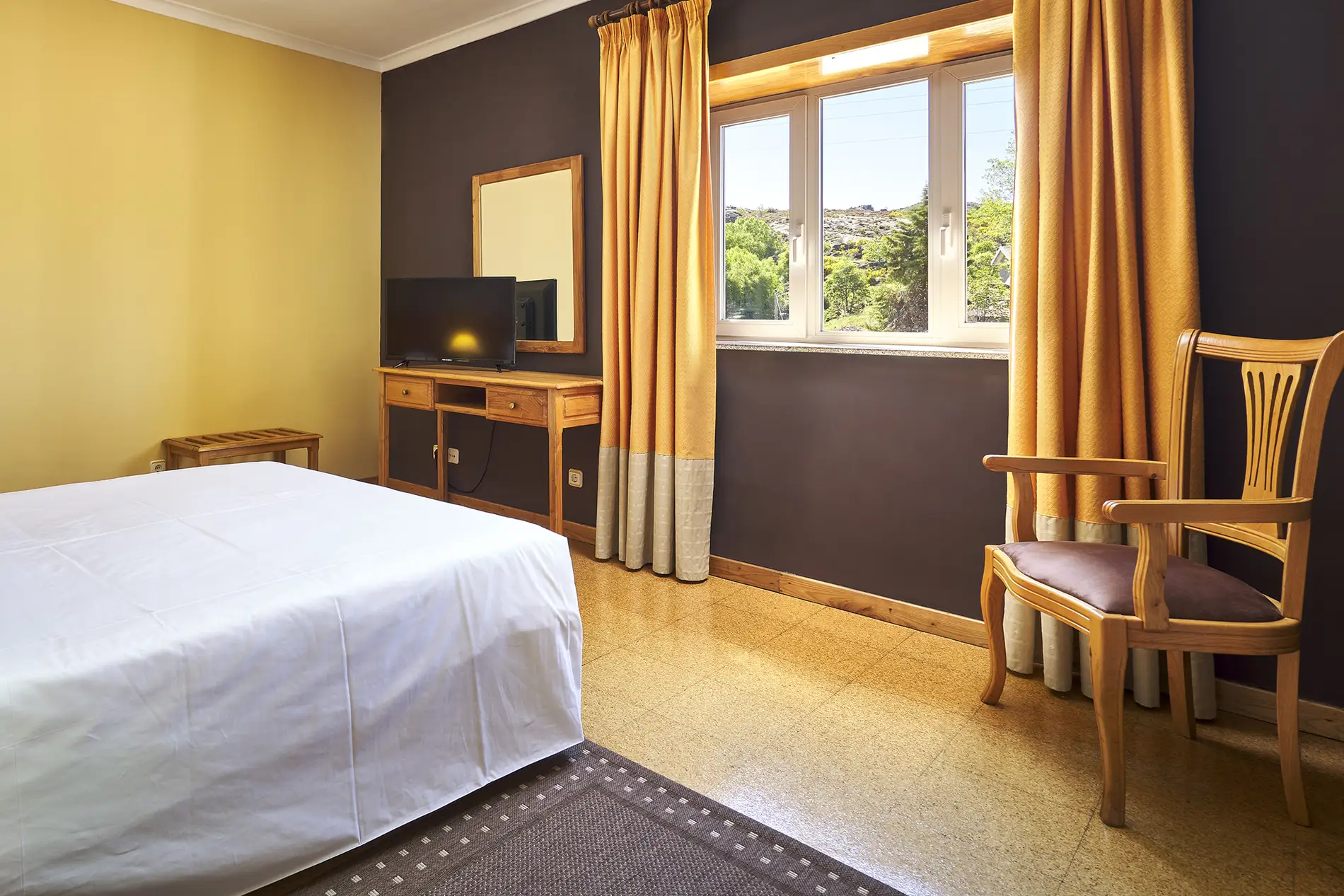 Quarto com cama, cortinas amarelas e TV perto de janela com vista verde