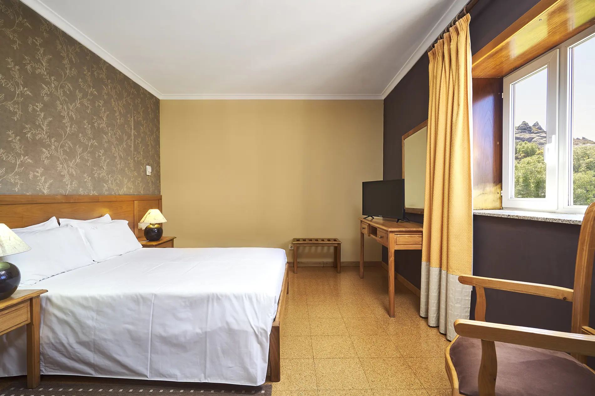 Quarto com cama, cortinas amarelas e TV, vista pela janela com montanhas ao fundo